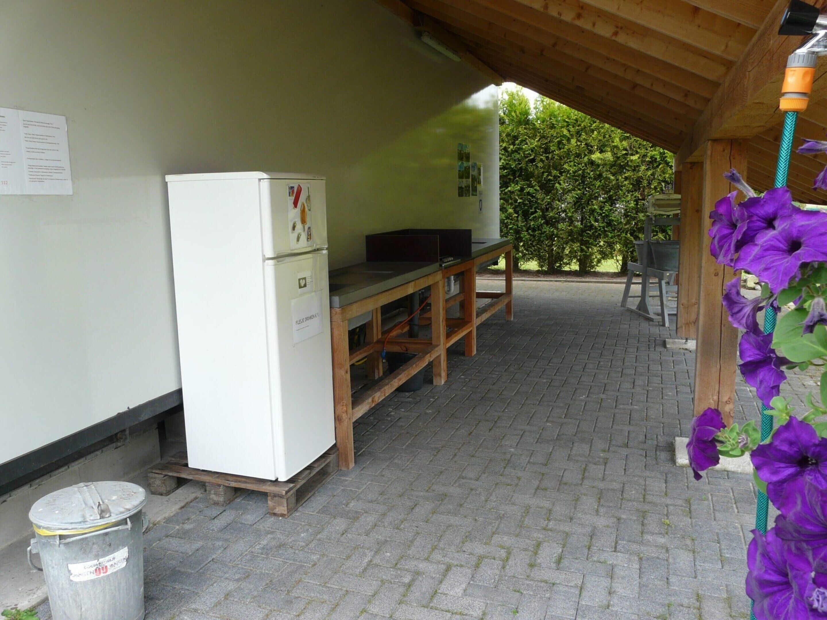 Un espace extérieur abrité comprenant un réfrigérateur blanc, une table en bois et une poubelle.