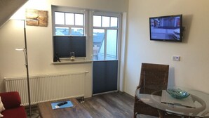 Una habitación con una mesa de madera, una silla de mimbre, un sofá rojo, una lámpara de pie, un televisor en la pared y una ventana con persianas.