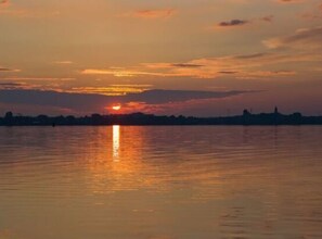 Auringonlasku tyynen vesistön yllä, taustalla kaupungin siluetti.