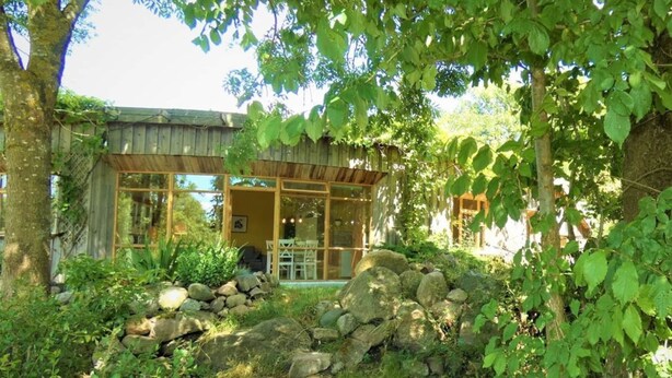 Un chalet rustique avec une façade en verre, entouré d’un mur en pierre naturelle et d’une végétation luxuriante.