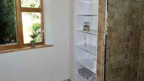 Une salle de bain avec une fenêtre, une étagère sur laquelle se trouvent un pot et une bougie, et un espace de douche carrelé.