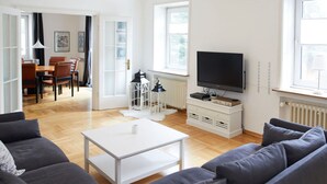 Una sala de estar moderna con un sofá gris, una mesa de centro blanca y un televisor sobre un mueble bajo. Se observa un comedor con una mesa y sillas al fondo.