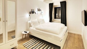 Un dormitorio con cama blanca, mesita de noche, lámpara, una alfombra pequeña y un armario.