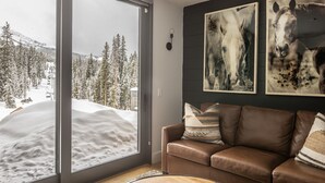 A living room with a large window overlooking a snowy mountain landscape, a brown leather sofa, and two horse paintings on the wall.