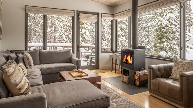 A modern living room with a large fireplace, grey sofas, and a view of snow-covered trees through large windows.