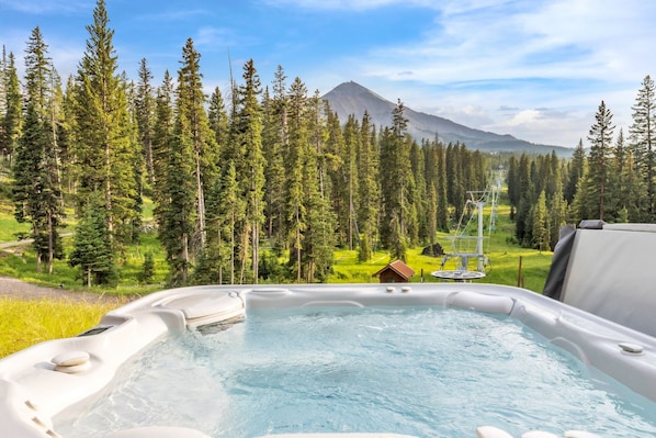 A hot tub with a mountain view, surrounded by a forest and a small cabin.
