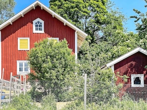 A red barn with a white window and a yellow shutter, surrounded by greenery and a chain-link fence. - 5 Person Holiday Home in Valdemarsvik (Valdemarsvik)