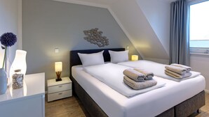 Une chambre d’hôtel moderne avec un grand lit, deux tables de chevet, une petite table, un élément décoratif mural et une fenêtre avec des rideaux.