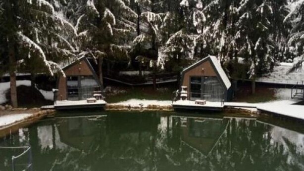 Dos cabañas separadas por un pequeño estanque, rodeadas de árboles cubiertos de nieve.