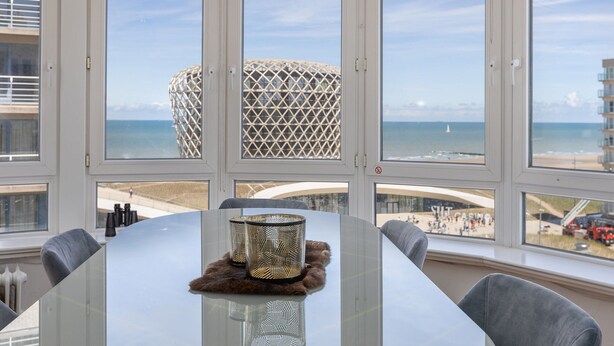 Een moderne eetruimte met een glazen tafel, grijze stoelen en uitzicht op het strand en de oceaan door grote ramen.