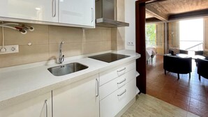 Een moderne keuken met witte kastjes, een roestvrijstalen spoelbak en een afzuigkap. Een eethoek met een tafel en stoelen is zichtbaar door een open deur.