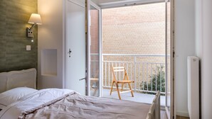 Une chambre à coucher avec un lit, une table de chevet, une lampe fixée au mur et une vue sur un bâtiment en briques, visible à travers une porte ouverte.