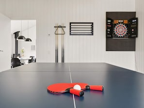 A ping pong table with a red paddle and ball, a dartboard, and a billiards table in a modern room with white walls and a dining area visible in the background. - 5 Star Holiday Home in Albaek (Aalbaek)