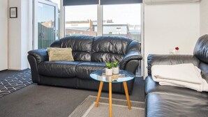 Een moderne woonkamer met een zwarte leren bank, een glazen salontafel en uitzicht op de stad door grote glazen deuren.