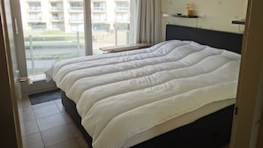 A hotel room with a large bed, a nightstand, a window with a view of a building, and a door to the outside.