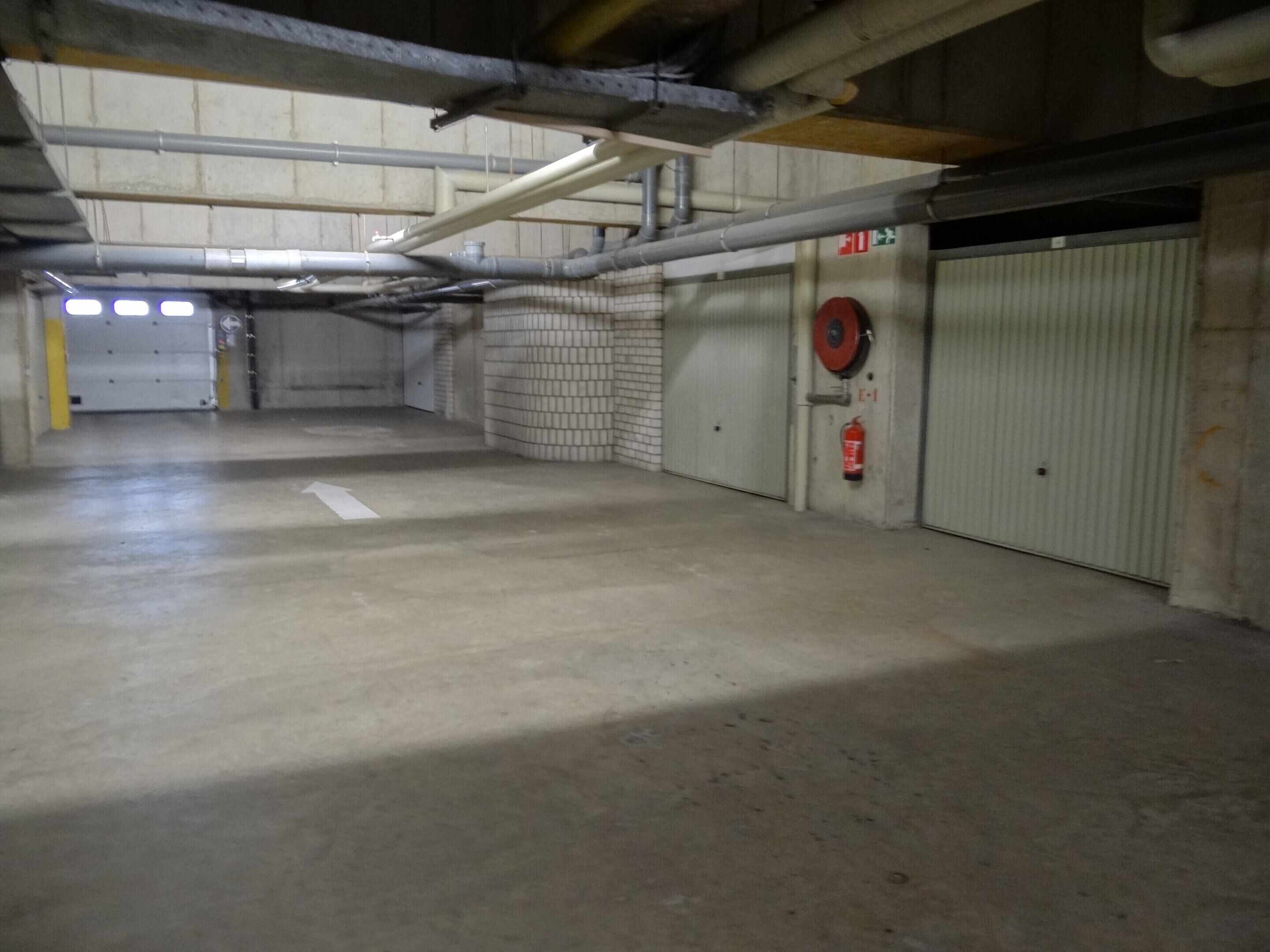 Un garaje estacionamiento interior con pisos y paredes de concreto, tuberías y conductos visibles, y un extintor de incendios montado en la pared.