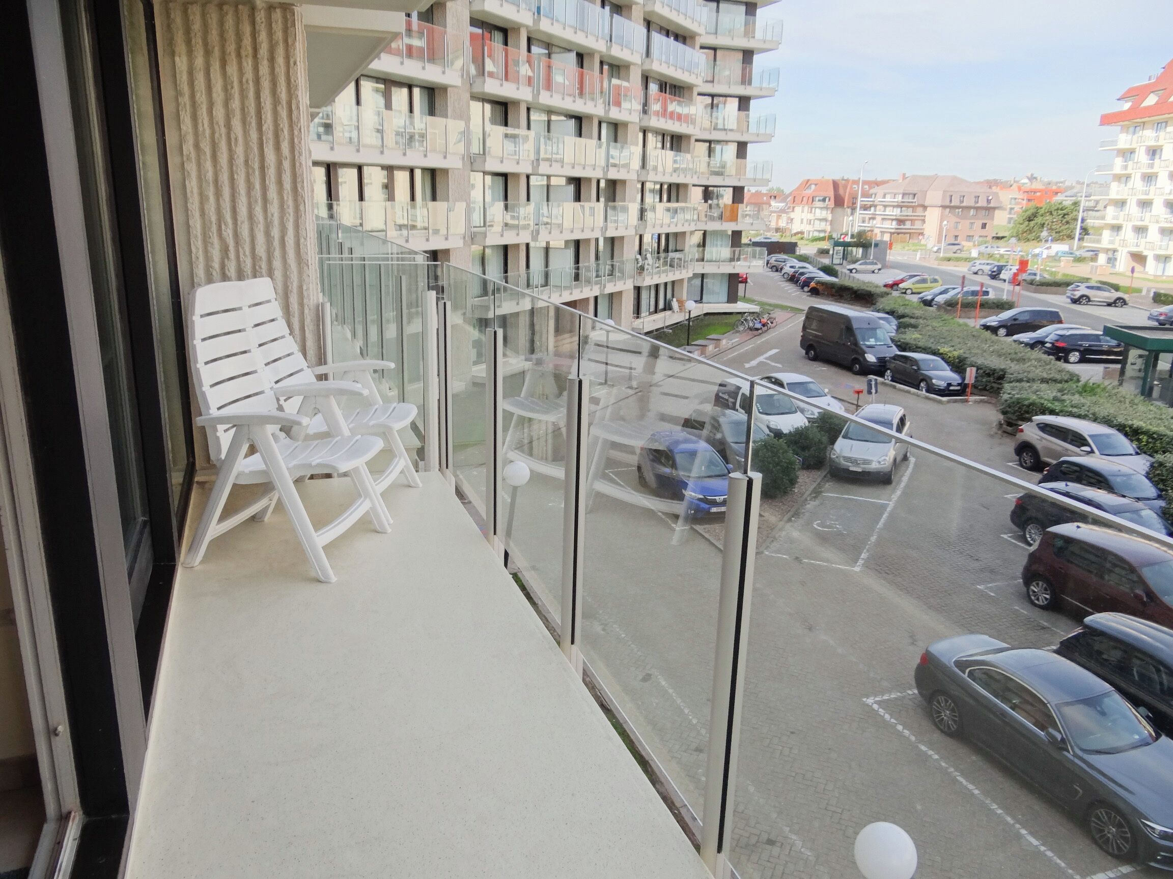 A balcony with white plastic chairs, a view of a parking lot with cars, and a multi-story building with balconies in the background.