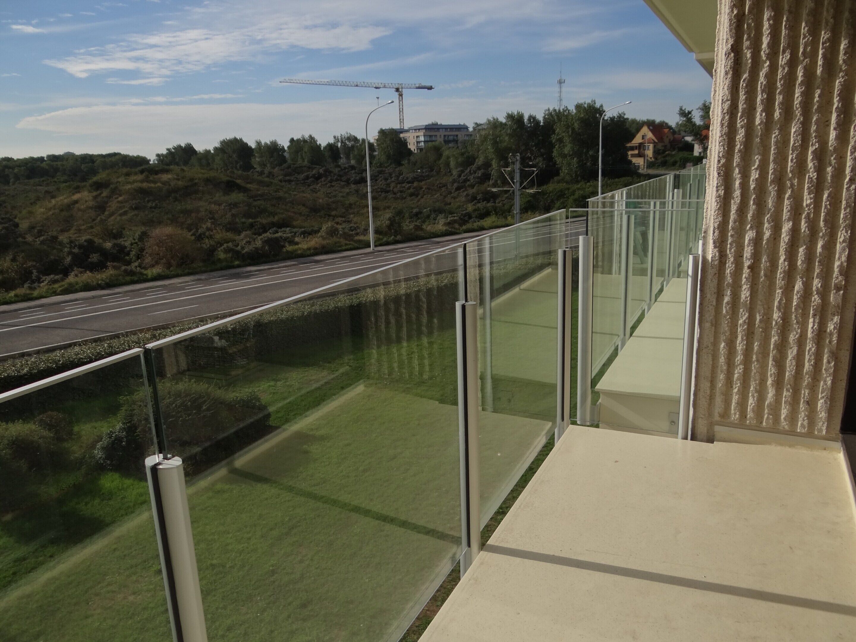 Un balcon doté d’une balustrade en verre, donnant sur une route et sur un espace verdoyant.