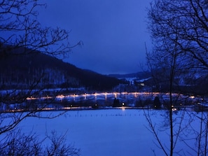 Une vue nocturne d’un lac où se reflètent les lumières, entouré d’arbres et de collines.