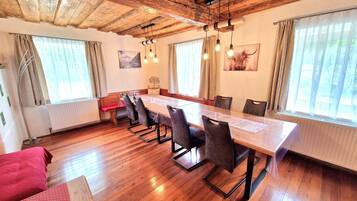 A dining area with a long wooden table, six chairs, and a bench. There are two windows with curtains, a radiator, and a wooden ceiling.