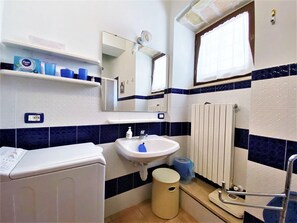 Une salle de bain avec un lavabo blanc, des murs recouverts de carreaux bleus et blancs, un miroir, une fenêtre avec un rideau et une petite poubelle.