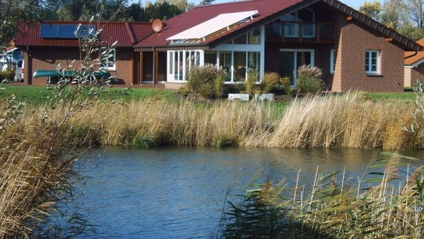 Une maison à deux étages avec un toit rouge, une grande véranda à l’avant et un étang au premier plan.