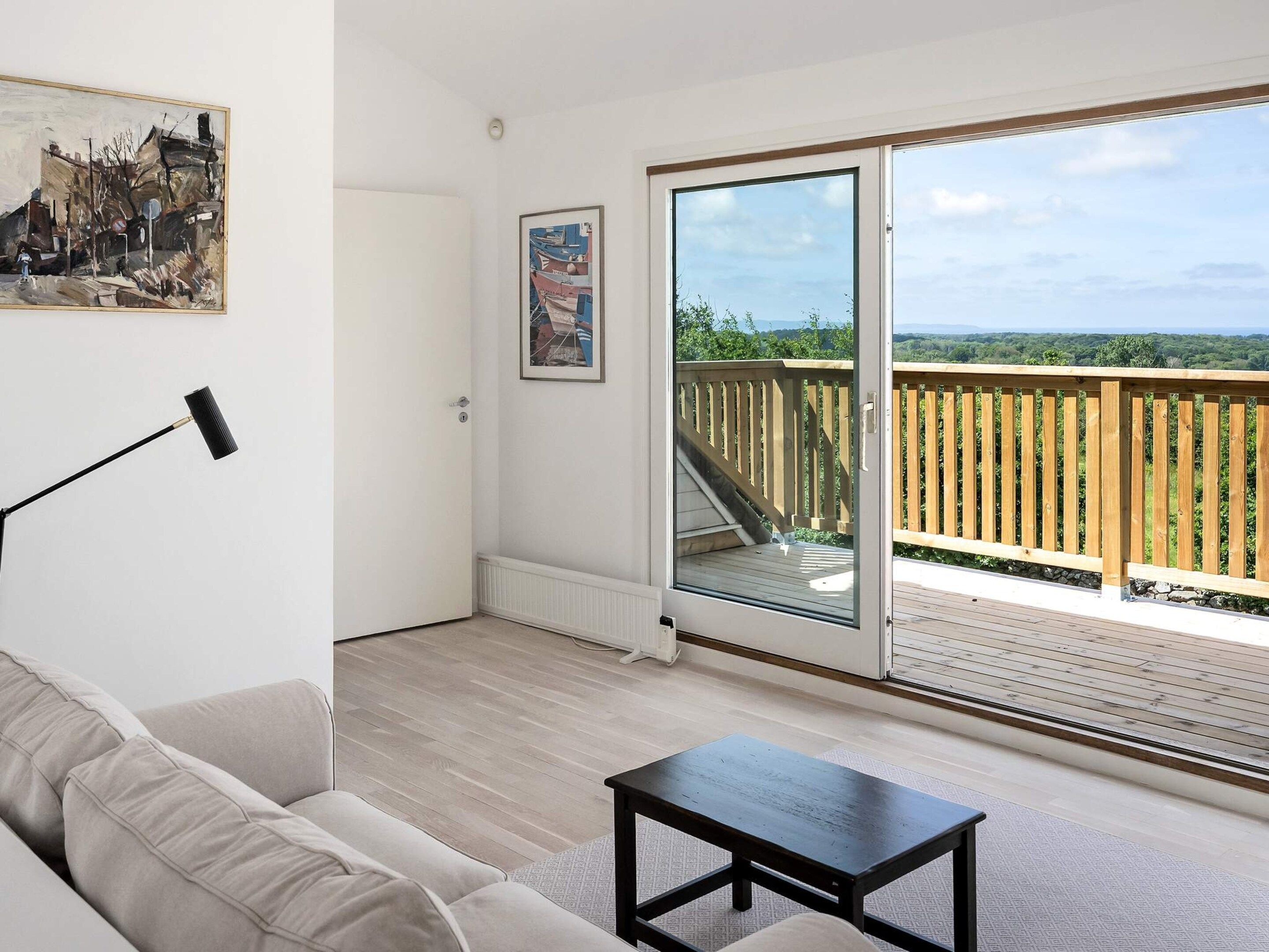 A room with a sofa, a small table, a painting, and a sliding glass door leading to a balcony with a wooden deck.