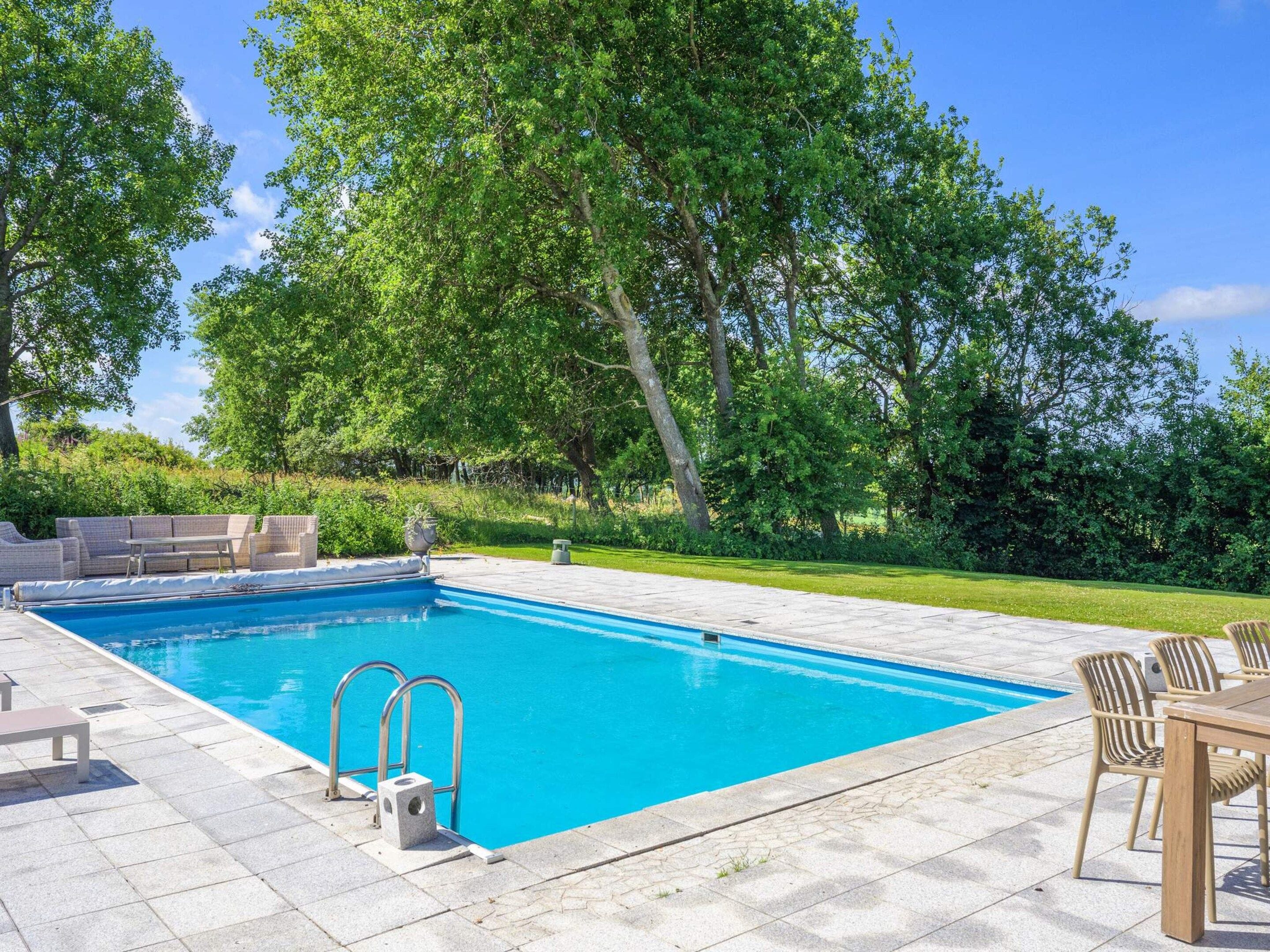 A rectangular swimming pool with a tiled deck, surrounded by lush greenery and trees.