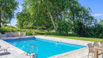 A rectangular swimming pool with a tiled deck, surrounded by lush greenery and trees.
