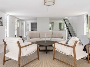 Een moderne woonkamer met een grote beige hoekbank, twee houten fauteuils, een ronde salontafel en een trap op de achtergrond.