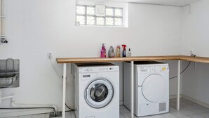 A laundry room with two white washing machines, a wooden shelf with cleaning supplies, and a window above the machines. - 5 Star Holiday Home in Bastad (Bastad)