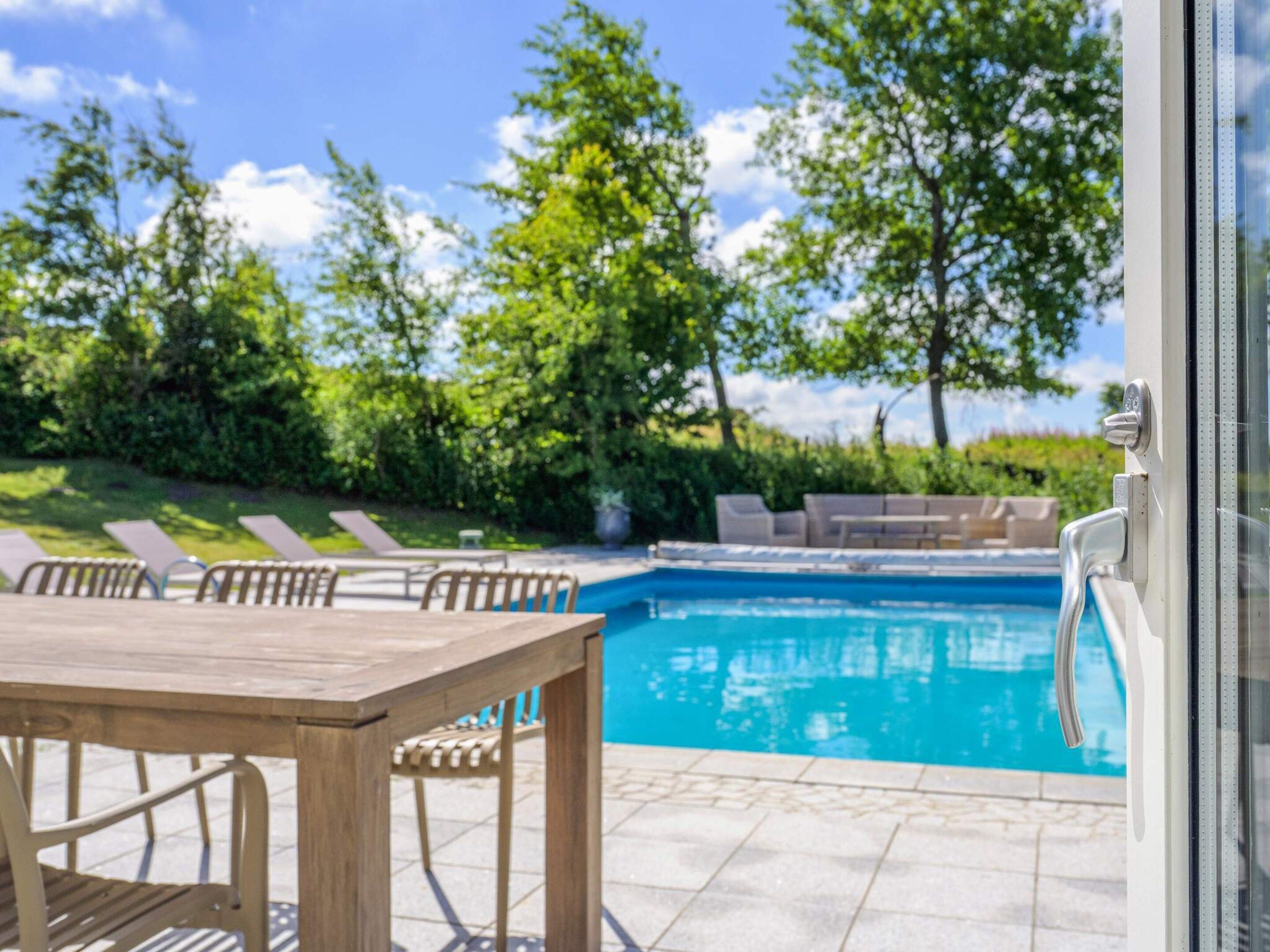 A pool area with a wooden table and chairs, a pool, and a view of trees and sky.