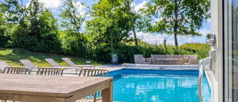 A pool area with a wooden table and chairs, a pool, and a view of trees and sky.