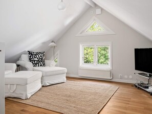 A modern, white-themed room with a sloped ceiling, a large window with a view of greenery, a white sofa, a small table, and a flat-screen TV. - 5 Star Holiday Home in Bastad (Bastad)