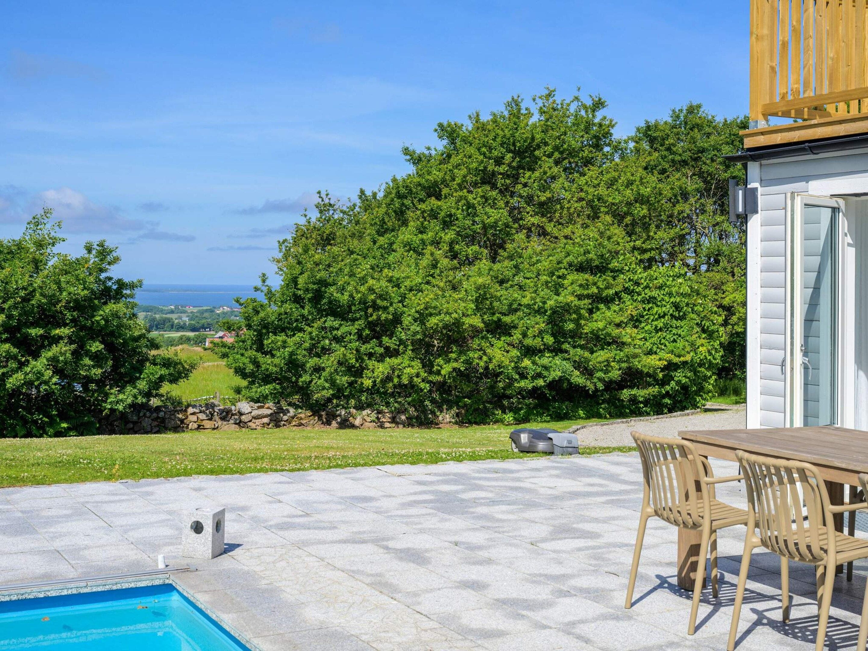 A pool with a stone deck, a wooden table with chairs, and a view of a green landscape and ocean.