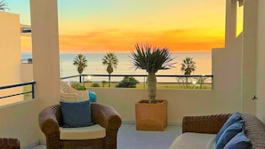 A balcony with wicker furniture, a potted plant, and a view of the sunset over the sea.
