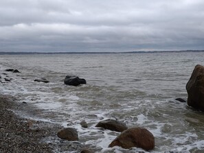 A rocky shoreline with waves, a distant city skyline, and overcast skies. - 6 Person Holiday Home in Knebel (Knebel)