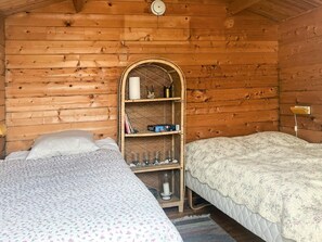 Acogedora habitación tipo cabaña con dos camas, una pared de madera, un estante con libros y velas, y un pequeño reloj.