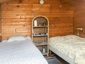 A cozy cabin room with two beds, a wooden wall, and a shelf with books and candles.