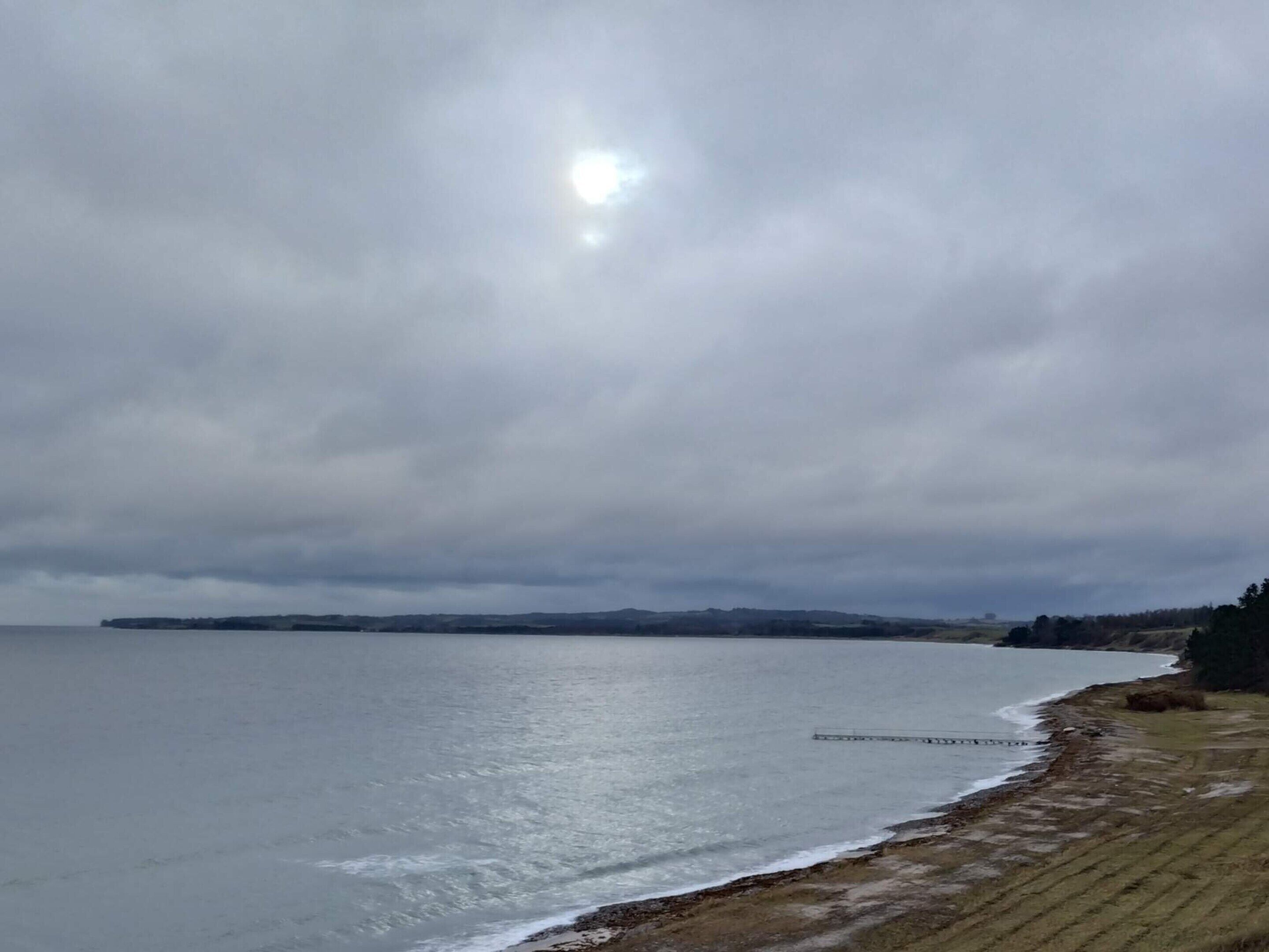 A coastal landscape with a body of water, a sandy beach, and distant hills under a cloudy sky.