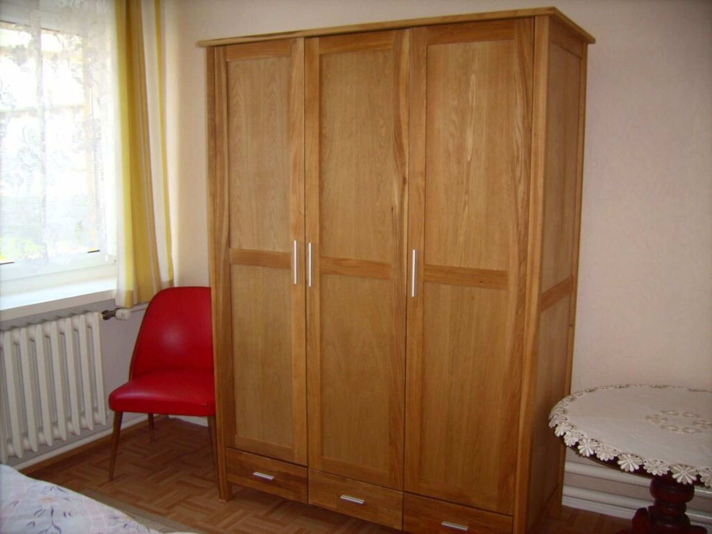 A room with a wooden wardrobe, a red chair, a small round table with a lace tablecloth, and a radiator.