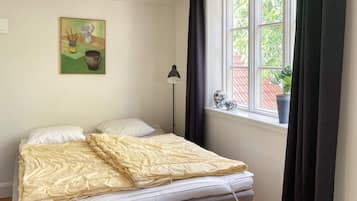 A bedroom with a bed, a bedside lamp, a window with curtains, and a painting on the wall.