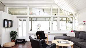 A modern living room with a sofa, a round coffee table, a black chair, a television mounted on the wall, and a large window with decorative items. - 5 Person Holiday Park Home in Karrebaeksminde (Karrebæksminde)