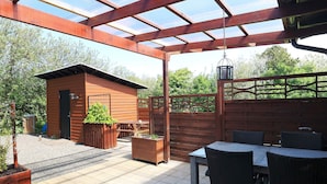 A covered patio with a wooden table and chairs, a small shed, and a garden area. - 6 Person Holiday Home in Vestervig (Vestervig)