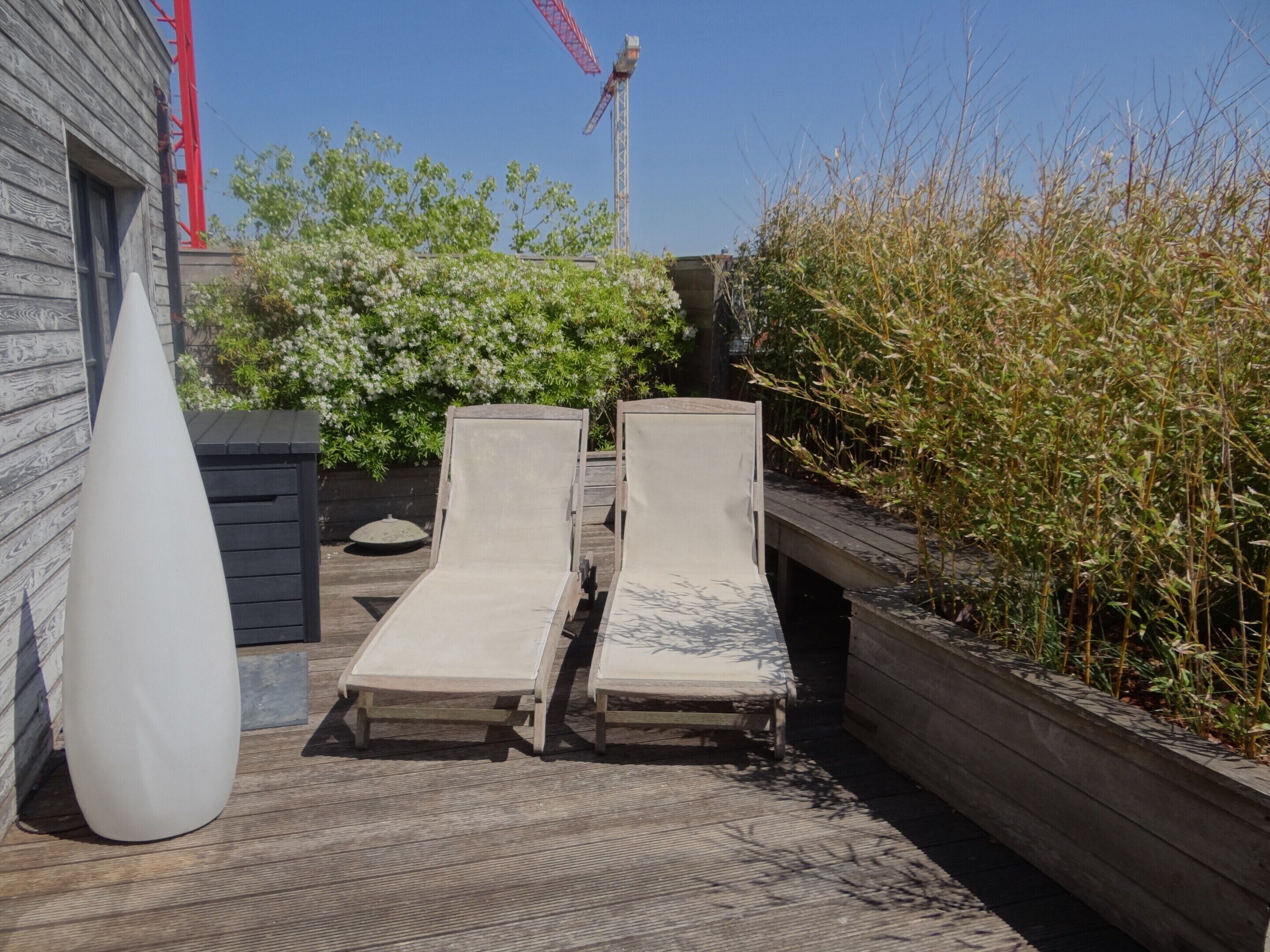 Deux fauteuils de jardin sur une terrasse en bois, avec un jardinière et un vase blanc.