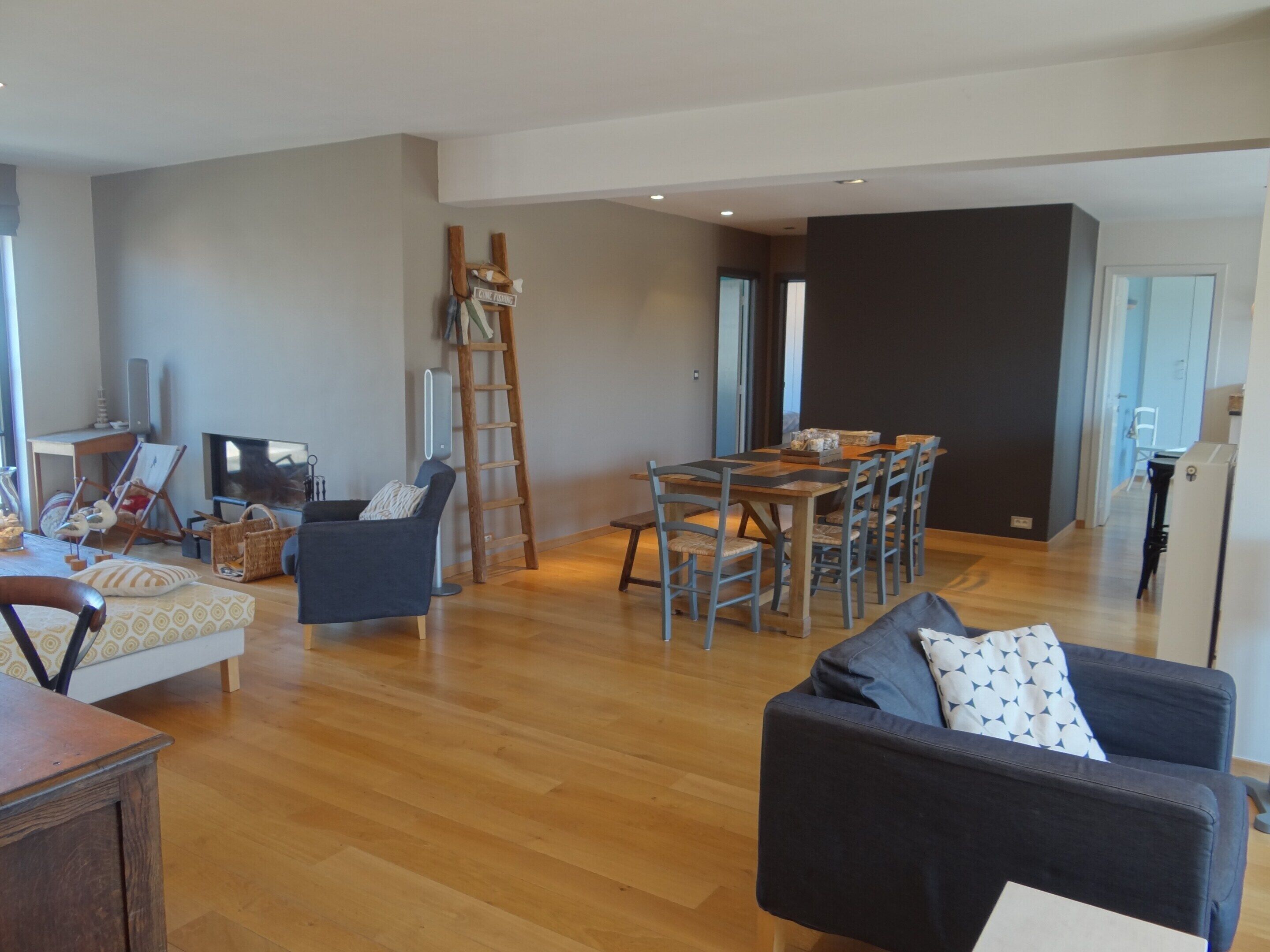 A spacious living area with a wooden floor, a dining table with chairs, a sofa, and a TV.