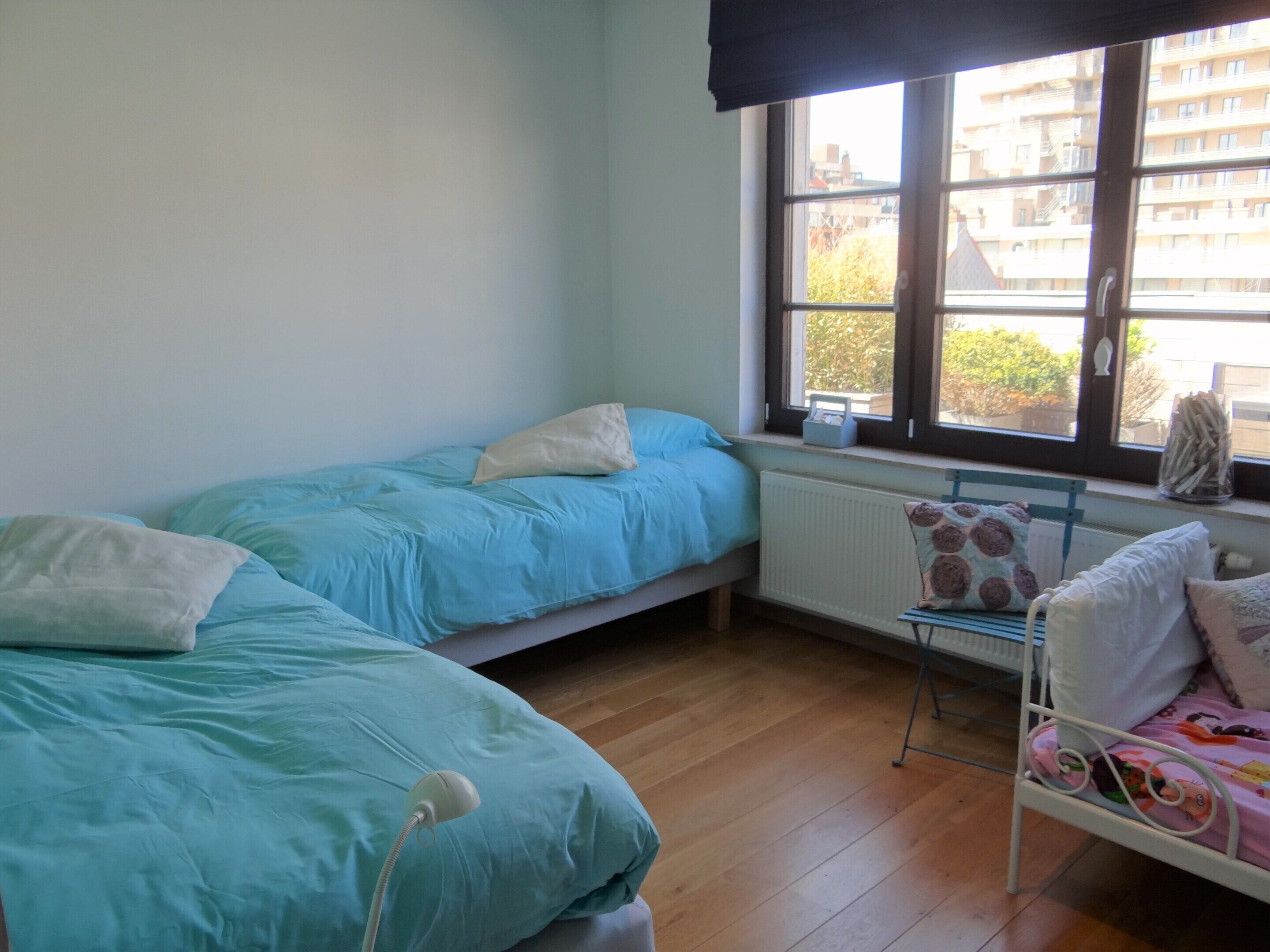 A dormitory room with two single beds, a radiator, a window with a view of buildings, and a chair with a cushion.