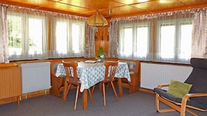 Una habitación acogedora con detalles de madera, una mesa de comedor con cuatro sillas, un sofá y ventanas amplias con cortinas.