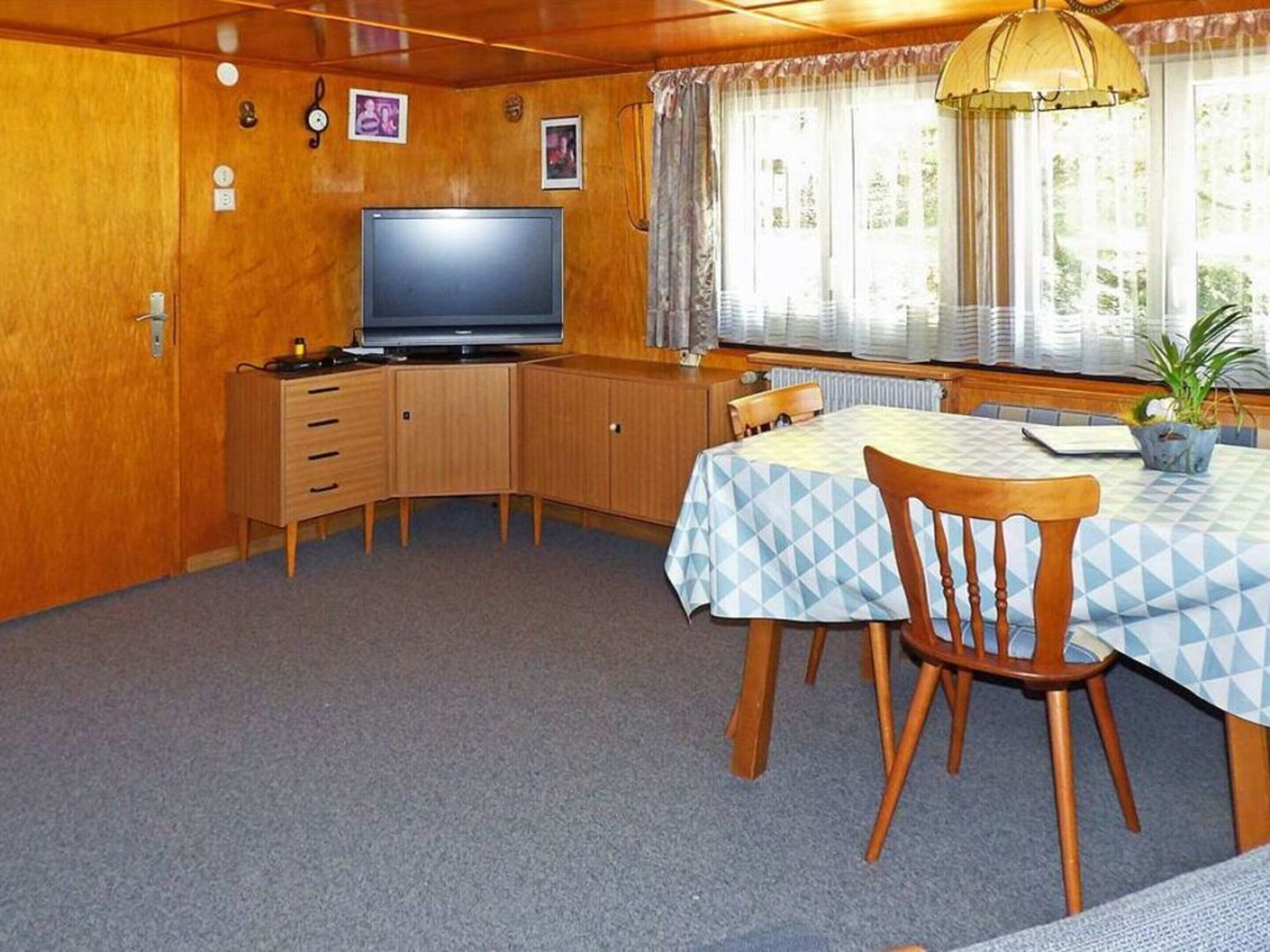 Una habitación pequeña con un armario de madera, un televisor, una mesa de comedor con sillas y una ventana con cortinas.
