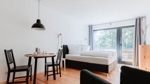 Ein modernes, minimalistisches Zimmer mit Holzboden, einem schwarzen Esstisch mit zwei Stühlen, einer schwarzen Couch und einem großen Fenster mit Blick auf Bäume.